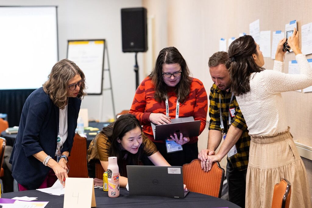 Several Code for America Summit attendees huddled around computers at a table and looking at sticky notes on a wall